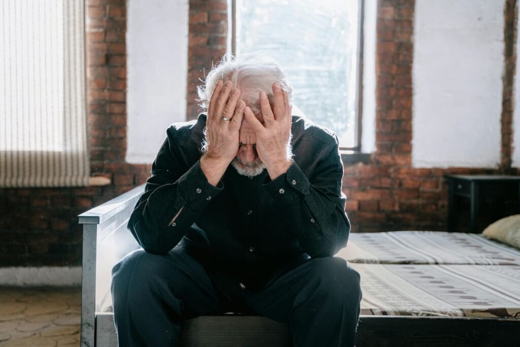 An older man sits on the edge of a bed, holding his head in his hands, visibly distressed.