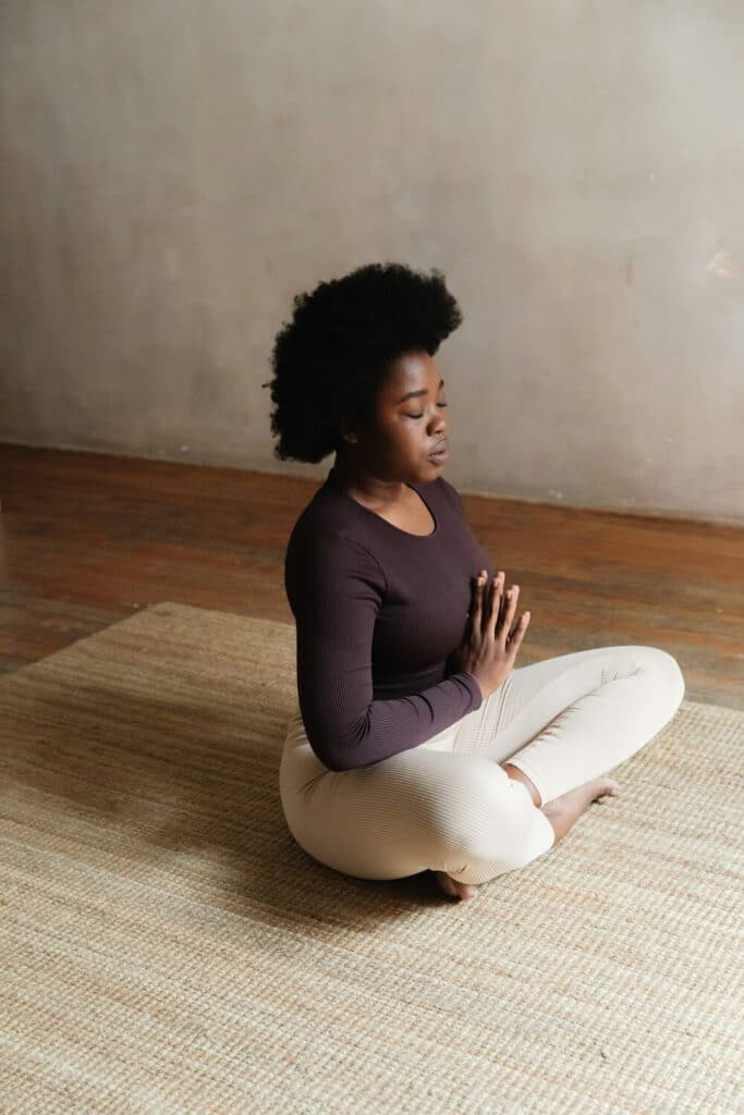 A woman with natural curly hair sits cross-legged on a woven mat, meditating with her hands in a prayer position.