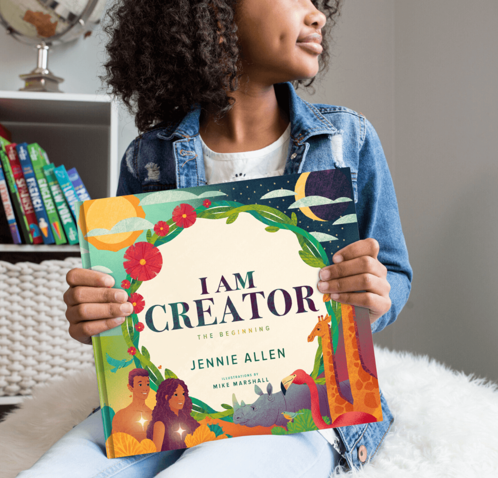 A young girl holding the children’s book "I Am Creator" by Jennie Allen, featuring a vibrant cover with animals and nature-inspired illustrations. She is seated in a cosy, well-lit room with a bookshelf in the background.