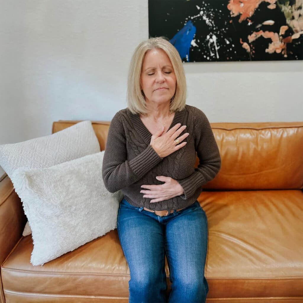 A woman sits calmly on a caramel-colored leather couch, her eyes gently closed as she practices mindful breathing. She places one hand on her chest and the other on her abdomen, symbolizing connection with her breath. She wears a dark brown knit sweater and blue jeans, exuding a relaxed and grounded energy. Soft white textured pillows are arranged next to her, adding comfort to the scene. Behind her, a modern abstract painting hangs on the light-colored wall, completing the tranquil and introspective atmosphere.