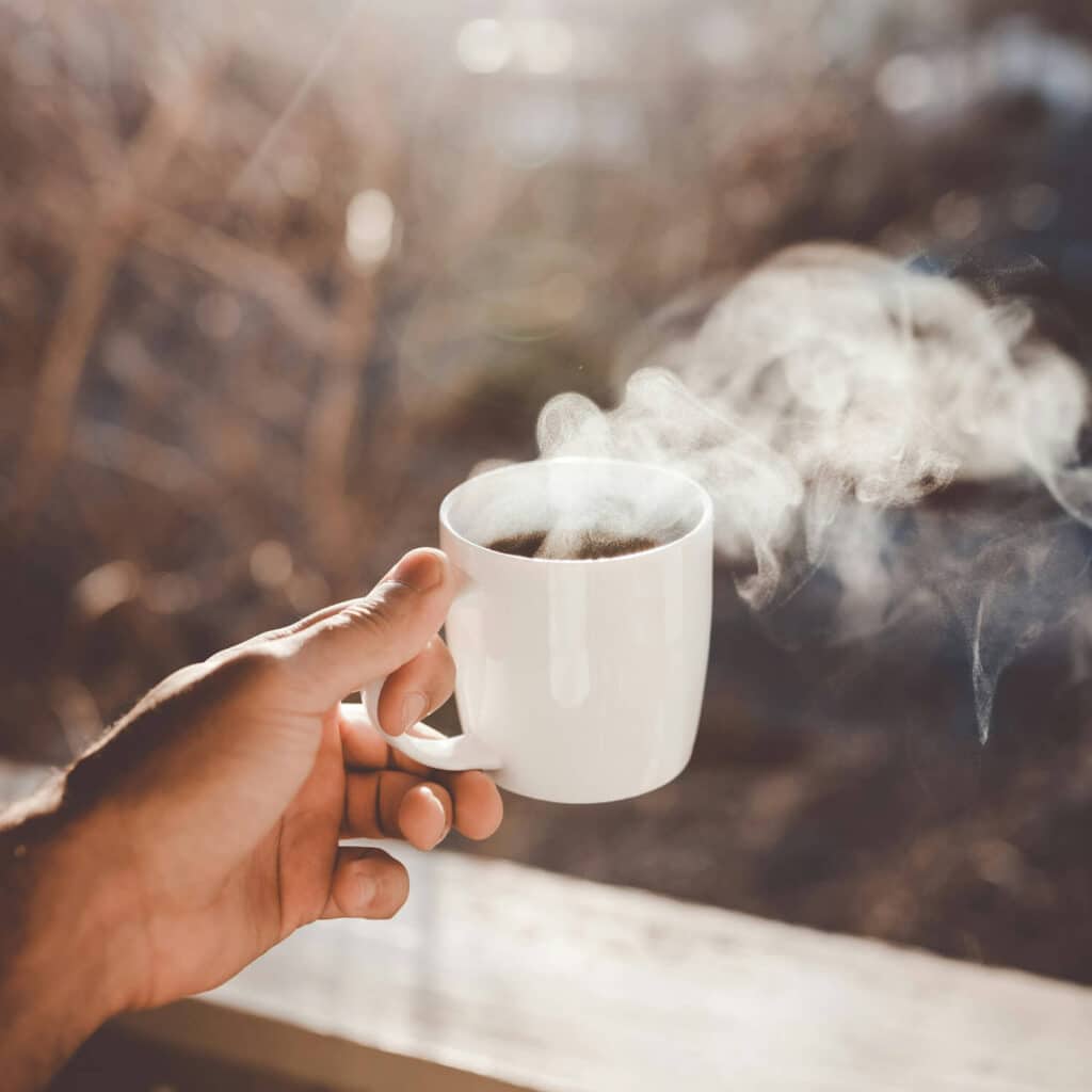 A hand holds a white mug filled with steaming coffee, with warm sunlight streaming in and a blurred outdoor background, evoking a peaceful and mindful morning.