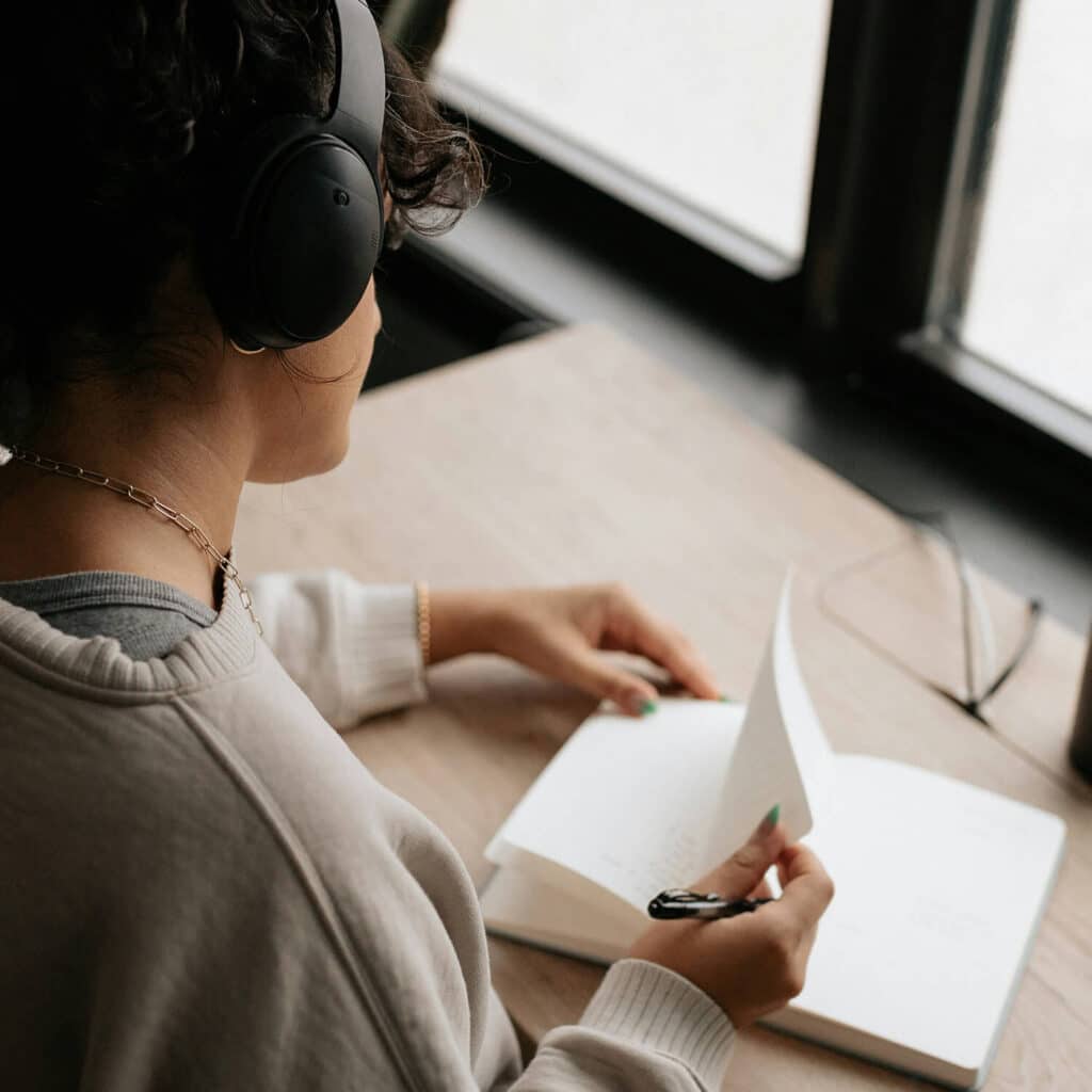 A person wearing black headphones sits at a wooden desk, flipping through the pages of a journal while holding a pen, creating a moment of reflection and focus.