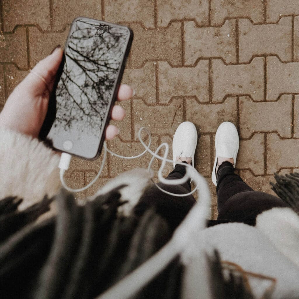 A person dressed in a cozy coat, black leggings, and white sneakers holds a phone with wired earbuds plugged in, reflecting the trees above in the phone screen while walking on a paved path.