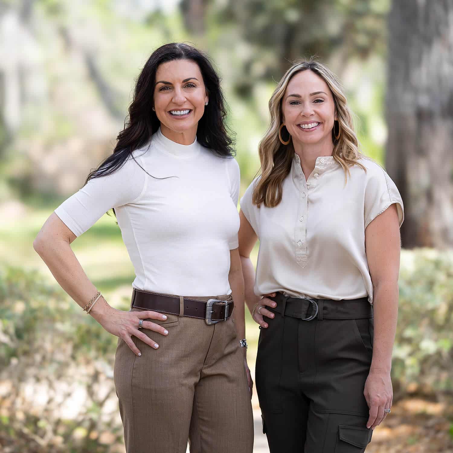 Jamie Schofield and Ashlee Seek, founders of Faithfully Guided Health Center, stand side by side outdoors in a sunlit, tree-lined setting. Both are smiling confidently at the camera. Jamie wears a fitted white top with brown trousers and a belt, while Ashlee wears a light beige blouse with dark pants. The background is softly blurred, highlighting a peaceful, natural environment.