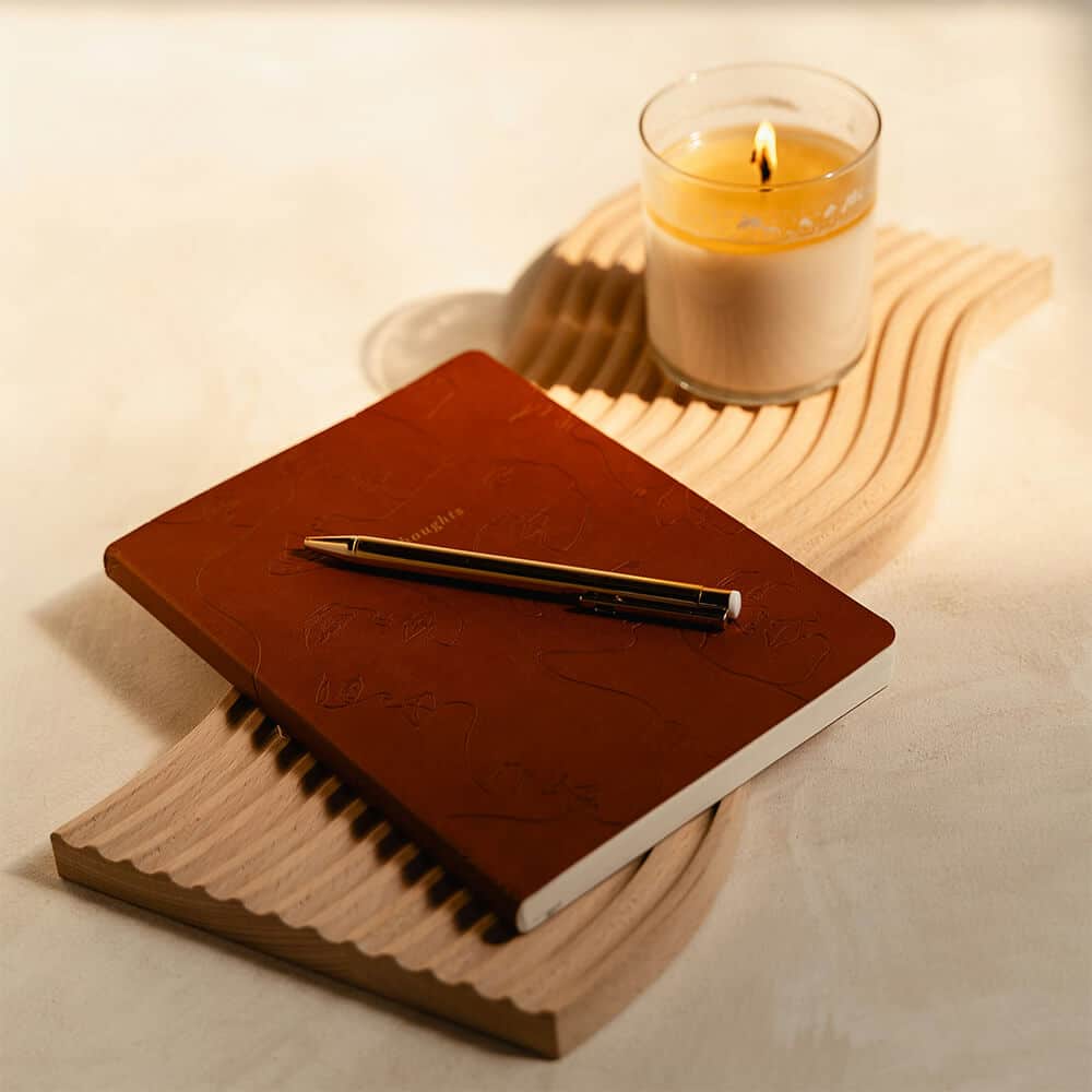 A brown leather journal and pen resting beside a lit candle on a sculpted wooden tray, evoking a calm, reflective atmosphere ideal for stress reduction.
