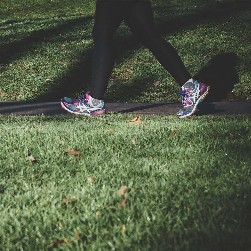 Close-up of a person’s legs in black leggings and colourful running shoes walking along a path through green grass, promoting the benefits of regular movement for autoimmune health.