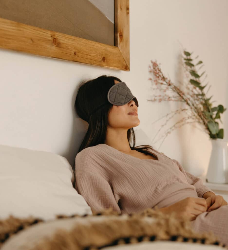 Middle-aged woman reclining in bed wearing a sleep mask, practicing relaxation for better sleep, with soft natural light and a calm bedroom setting.