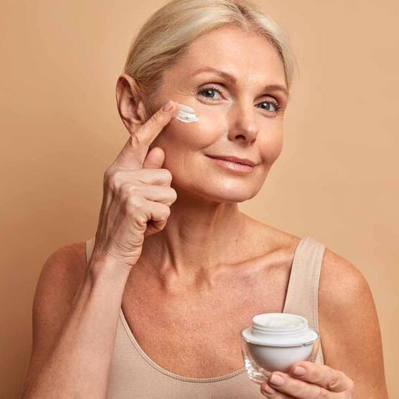 A mature woman applying face cream to her cheek while holding a small jar, standing against a neutral beige background.